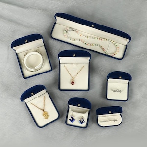 May include: A collection of navy blue velvet jewellery boxes in various shapes and sizes, each lined with cream-coloured fabric. The boxes contain a pearl necklace, a silver bracelet, a gold pendant necklace, a ring, and blue crystal earrings.
