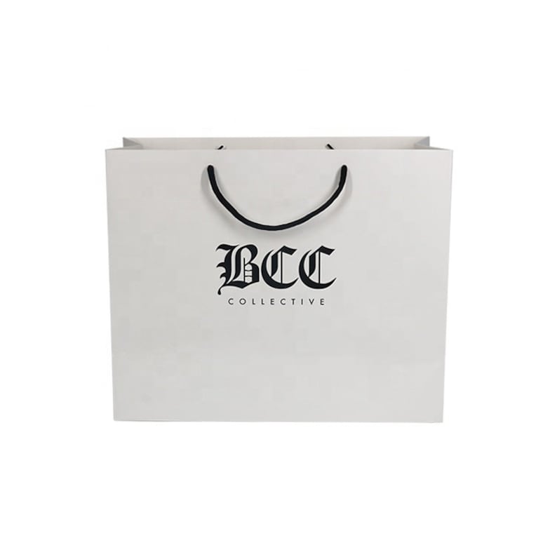 May include: A white shopping bag with black rope handles. The bag features the text "BCC COLLECTIVE" in a gothic font. The bag is rectangular and has a clean, minimalist design, suitable for retail or gift packaging.