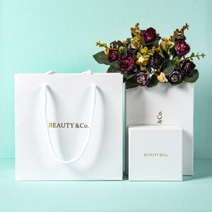May include: White gift bag, box, and shopping bag with the gold text "BEAUTY & Co." A bouquet of dark purple flowers sits atop the shopping bag. The gift bag has white rope handles.