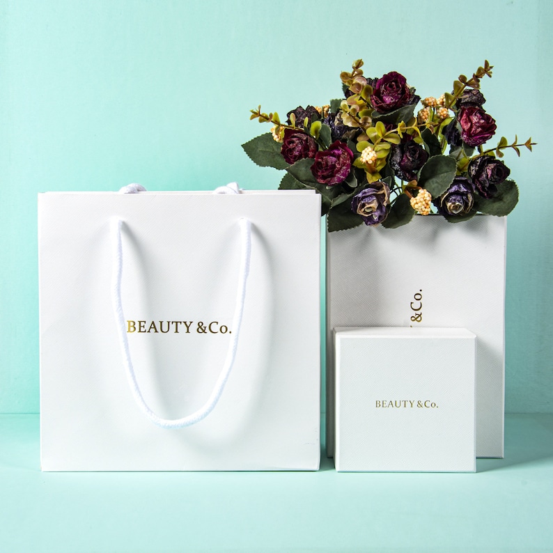 May include: White gift bag, box, and shopping bag with the gold text "BEAUTY & Co." A bouquet of dark purple flowers sits atop the shopping bag. The gift bag has white rope handles.