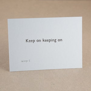 以下が含まれることがあります： 「Keep on keeping on」というフレーズが黒で印刷された白いカード。カードはわずかに傾いており、端が見えています。アーティストのサイン「Georgu E」が下部にあります。
