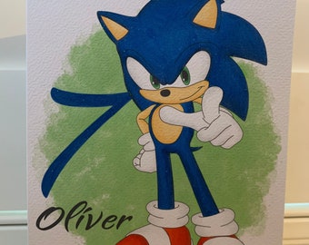 May include: A birthday card featuring a blue cartoon hedgehog character with red shoes, giving a thumbs-up gesture. The card has the text "Happy Birthday Oliver" in black script, set against a green watercolor-style background.