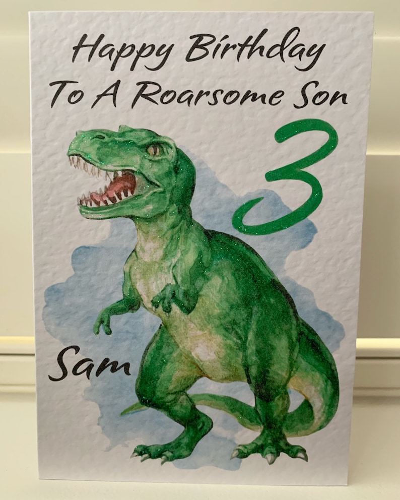 May include: A birthday card with a watercolor illustration of a green Tyrannosaurus Rex. The card reads "Happy Birthday To A Roarsome Son" with the number "3" and the name "Sam". The dinosaur has glitter accents.