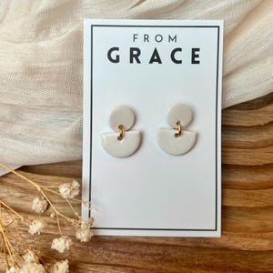 Puede incluir: Pendientes de cerámica blanca con un diseño geométrico. Los pendientes están en una tarjeta blanca con el texto "FROM GRACE" impreso en ella.