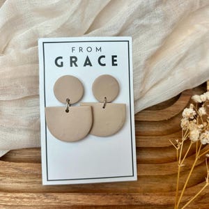 Puede incluir: Un par de pendientes de arcilla beige con un diseño de medio círculo y círculo. Los pendientes se muestran en una tarjeta blanca con el texto "FROM GRACE".