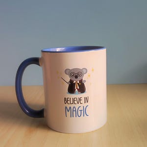 Op de afbeelding: Witte keramische mok met een blauw handvat en rand. De mok heeft een cartoon koala met een bril en een tovenaarsgewaad, die een toverstaf vasthoudt. De tekst "BELIEVE IN MAGIC" staat eronder. Op een houten oppervlak.