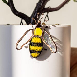 May include: A white ceramic planter with a small, yellow and black stained glass bee attached to the side. The planter is filled with a small green artificial plant.