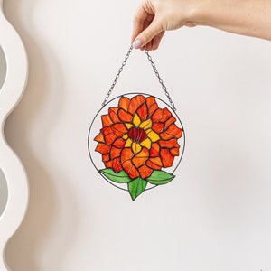 以下が含まれることがあります： 赤と黄色の花と緑の葉が描かれたステンドグラスのサンキャッチャー。サンキャッチャーはチェーンで吊り下げられており、白い壁に飾られています。