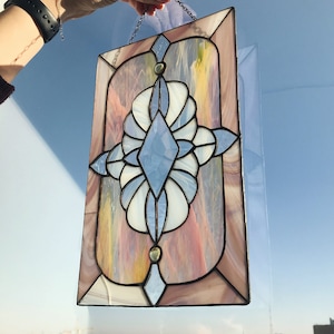 Puede incluir: Una ventana de vitral colgando de una cadena. La ventana es rectangular y presenta un diseño geométrico con un diamante azul en el centro. El vidrio es iridiscente y tiene una gama de colores rosa, amarillo y azul.