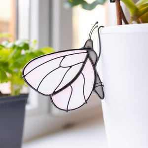 Pode incluir: Um ornamento de borboleta em vitral rosa pálido e branco com linhas de solda de metal escuro. A borboleta apresenta segmentos de asa delicados e duas antenas finas, projetada para prender na borda de um vaso de plantas cilíndrico branco.