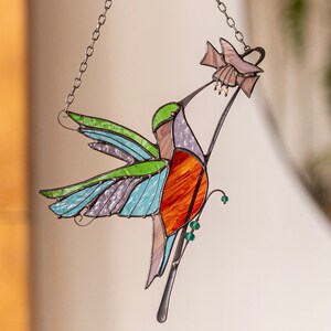 Op de afbeelding: Een gekleurde glazen kolibrie met groene, blauwe en oranje vleugels, die op een tak zit met een bloem. De kolibrie hangt aan een zilveren ketting.
