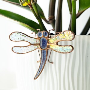 May include: A dragonfly brooch with iridescent wings and a blue body, set against a white background with green plant stems and leaves. The dragonfly is made of glass and metal.