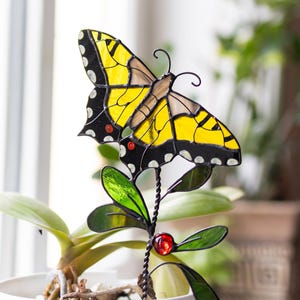 Könnte beinhalten: Ein Schmetterlingsornament aus Buntglas mit leuchtend gelben Flügeln, schwarzen Rändern, weißen Punkten und kleinen roten Akzenten. Der Schmetterling ist an einem gedrehten Metallstab mit grünen Glasblättern und einer roten Glasperle befestigt.