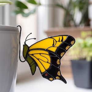 May include: A stained glass butterfly ornament featuring vibrant yellow and black wings with small gold accents and a single red dot. The butterfly is attached to a black metal hook designed to hang securely over the edge of a plant pot.