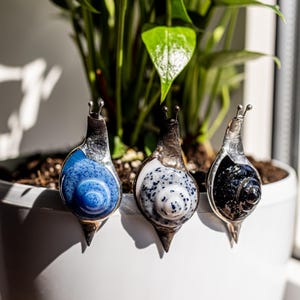 Puede incluir: Tres figuras decorativas de caracoles con conchas de vidrio en azul, blanco y negro, cada una con un cuerpo y antenas plateadas. Los caracoles están colocados en el borde de una maceta blanca, con follaje verde de fondo.