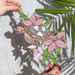 Op de afbeelding: Een glas-in-lood schildpad met een roze en groen bloemmotief. De schildpad is omringd door bloemen en bladeren, en het glas is iriserend.