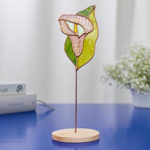 May include: A stained glass calla lily sculpture with a light pink and yellow flower and green leaf. The flower is mounted on a copper wire stem and a round wooden base. The sculpture is displayed on a blue surface.