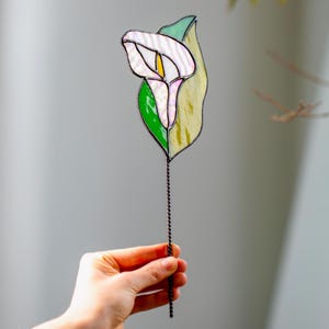 May include: A stained glass calla lily flower with a white bloom, yellow center, and green and yellow leaves. The flower is attached to a twisted metal stem. The flower is held by a hand.