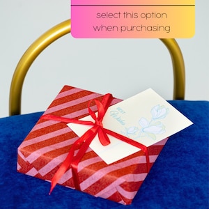 May include: A red and pink striped gift box with a red ribbon and a white card that says "Best Wishes" with a blue flower illustration. The box is sitting on a blue surface.