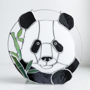 Pode incluir: Um panda de vidro colorido, com pelo preto e branco, segurando um caule de bambu verde na boca. O panda é rodeado por uma moldura de metal prateada.