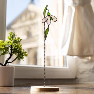 Può includere: Un fiore di bucaneve in vetro colorato su un supporto in filo di ferro nero con una base in legno. Il fiore è bianco con foglie verdi.