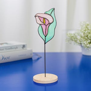 Könnte beinhalten: Eine Glasmalerei-Calla-Lilien-Skulptur. Die Blume ist rosa mit gelbem Zentrum, vor einem grünen Blatt. Sie steht auf einem Holzsockel, getragen von einem schwarzen Metallstiel. Die Skulptur ist ein dekorativer Artikel.