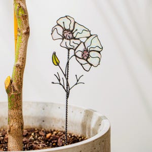 May include: A decorative metal flower stake featuring two white and pink stained glass flowers. The stake has a twisted stem and small branches, and is designed to be placed in a potted plant. The flower petals have a pearlescent sheen.