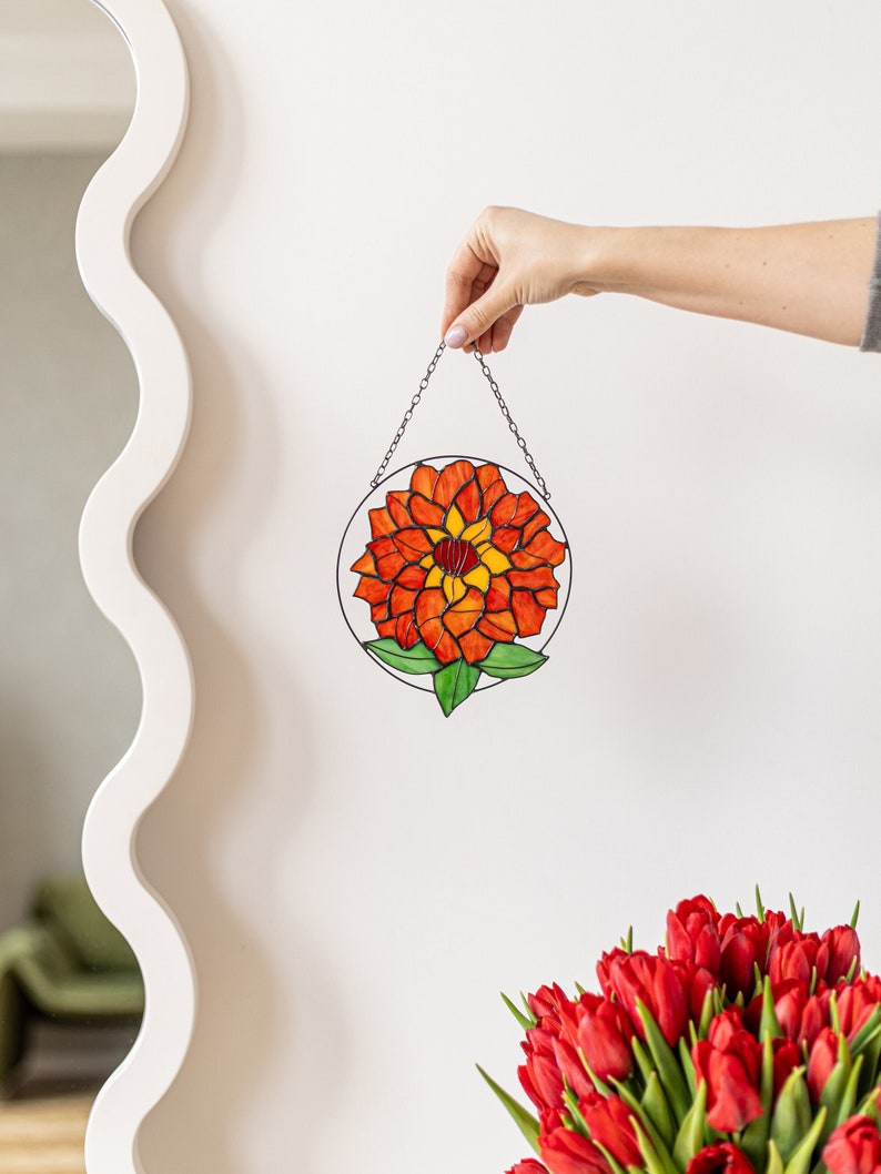 以下が含まれることがあります： 赤と黄色の花と緑の葉が描かれたステンドグラスのサンキャッチャー。サンキャッチャーはチェーンで吊り下げられており、白い壁に飾られています。
