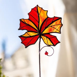 May include: A stained glass maple leaf garden stake. The leaf features vibrant red and yellow hues, with a black metal frame and stem. A small red bead accents the curled design at the bottom of the stake. The leaf measures approximately 8 inches in width.