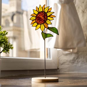 May include: A stained glass sunflower with yellow, orange, and red petals, green leaves, and a twisted wire stem. The sunflower is mounted on a round wooden base, creating a decorative accent piece.