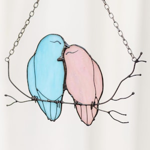 Puede incluir: Dos pájaros de vidrio coloreado, uno azul y otro rosa, están posados en una rama de alambre. Los pájaros se miran y parecen besarse.