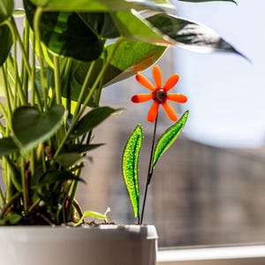 Peut inclure: Une décoration florale en verre composée de pétales orange vif avec un centre sombre, fixée sur une fine tige métallique avec deux feuilles en verre vert translucide et texturé, placée dans une plante verte en pot dans un cache-pot blanc.