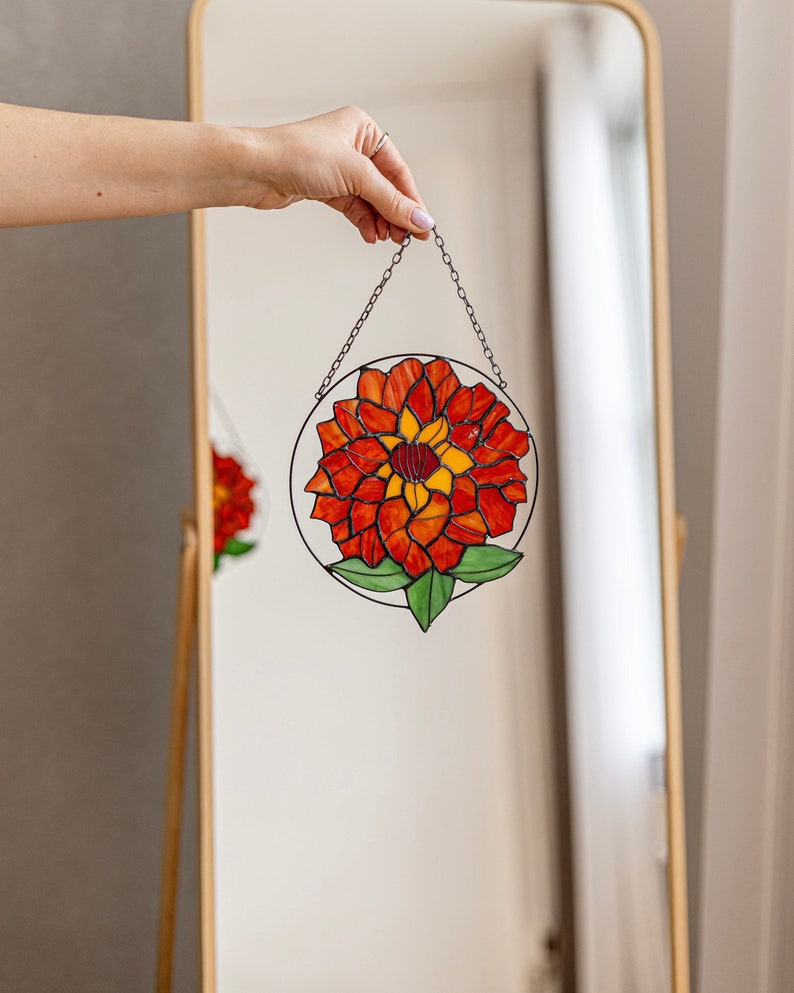 以下が含まれることがあります： 赤とオレンジの花と緑の葉が描かれたステンドグラスのサンキャッチャー。サンキャッチャーは丸く、黒いチェーンで吊り下げられています。
