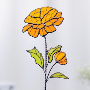 Pode incluir: Uma escultura de flor em vitral com uma grande flor laranja e um botão menor. A flor tem folhas verdes e uma haste de metal torcida. O vidro é de uma cor laranja vibrante.
