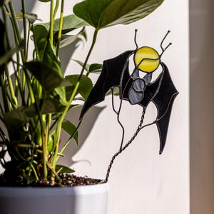 May include: A black stained glass bat with a yellow moon and gray body, attached to a twisted wire stake. The bat is placed in a white pot with a green plant.