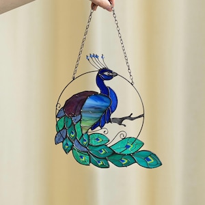 Puede incluir: Un atrapasol de vidrio coloreado con un diseño de pavo real azul y verde. El pavo real está posado en una rama con sus plumas de la cola extendidas. El atrapasol está colgado de una cadena.