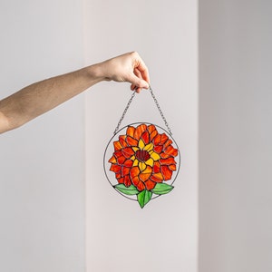 以下が含まれることがあります： 赤とオレンジの花と緑の葉が描かれたステンドグラスのサンキャッチャー。サンキャッチャーは銀色のチェーンで吊り下げられています。