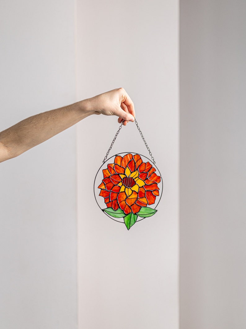 以下が含まれることがあります： 赤とオレンジの花と緑の葉が描かれたステンドグラスのサンキャッチャー。サンキャッチャーは銀色のチェーンで吊り下げられています。
