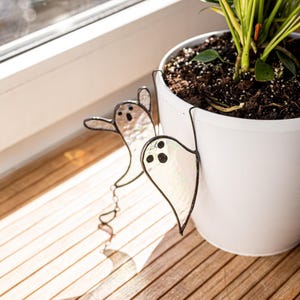 以下が含まれることがあります： 黒い縁取りのある、虹色のガラス製のゴーストプランターピックが2つ、白い植木鉢に取り付けられています。 ゴーストはシンプルな顔の特徴があり、鉢からぶら下がっています。 植木鉢には土と緑の植物が入っています。