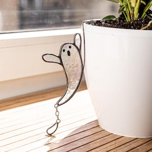 Puede incluir: Decoración de fantasma de vidriera nacarada, con contornos y rasgos faciales negros, colgando de una maceta blanca. El fantasma tiene una cadena y un lazo. La maceta está sobre una superficie de madera.