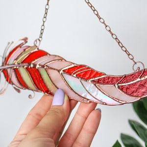 Peut inclure: Un attrape-soleil en verre coloré en forme de plume avec des pièces de verre rouge, rose et vert. La plume est suspendue à une chaîne en cuivre.