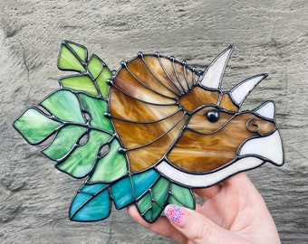 May include: A stained glass window hanging features a brown and white triceratops head with green leaves surrounding it.