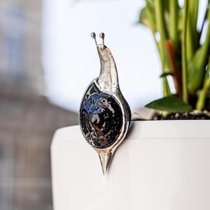 May include: A silver snail ornament with a shiny black shell and two tiny antennae. The snail is positioned on the edge of a white plant pot, with green leaves in the background. The figurine has a pointed base.