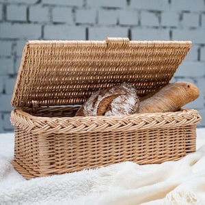Puede incluir: Una cesta de pan de mimbre con tapa, abierta para revelar dos panes. La cesta es de color marrón claro y tiene un patrón tejido. El pan es de color marrón claro y tiene una textura crujiente.