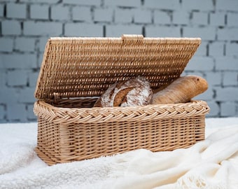 May include: A wicker bread basket with a lid, open to reveal two loaves of bread. The basket is light brown and has a woven pattern. The bread is a light brown color and has a crusty texture.