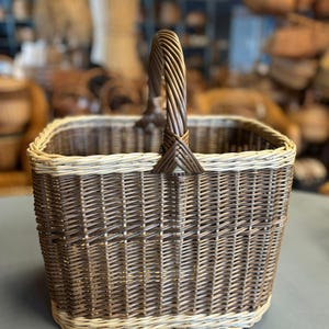 May include: A square wicker basket with a woven handle. The basket is primarily brown with a lighter tan border at the base and top. The handle is a twisted design. The basket is sitting on a gray surface.