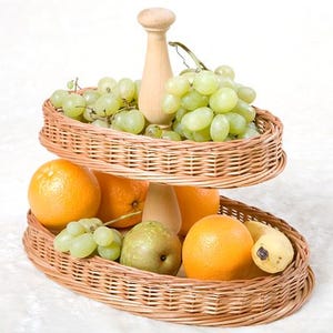 May include: Two-tiered wicker fruit basket with a wooden handle. The top tier holds green grapes and the bottom tier holds oranges, pears, and bananas.