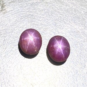Könnte beinhalten: Zwei ovale Sternrubin-Edelsteine. Die Edelsteine haben eine tiefrot-violette Farbe mit einem sechsstrahligen Sternmuster auf der Oberfläche. Die Steine sind glatt und poliert und reflektieren das Licht. Geeignet für die Schmuckherstellung.