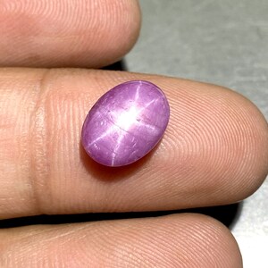 Könnte beinhalten: Ein ovaler Sternrubin-Edelstein im Cabochon-Schliff mit einem leuchtenden Magentarosa-Farbton. Der Stein weist einen ausgeprägten sechsstrahligen Asterismus-Effekt auf, bei dem ein helles weißes Sternmuster mittig auf der polierten Oberfläche erscheint.