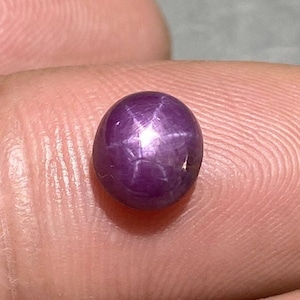 Könnte beinhalten: Ein runder, im Cabochon-Schliff gefertigter Sternrubin-Edelstein mit einem tiefen violett-roten Farbton. Der Stein zeigt einen ausgeprägten sechsstrahligen Sterneffekt, bekannt als Asterismus, als helles, sich kreuzendes weißes Lichtmuster.